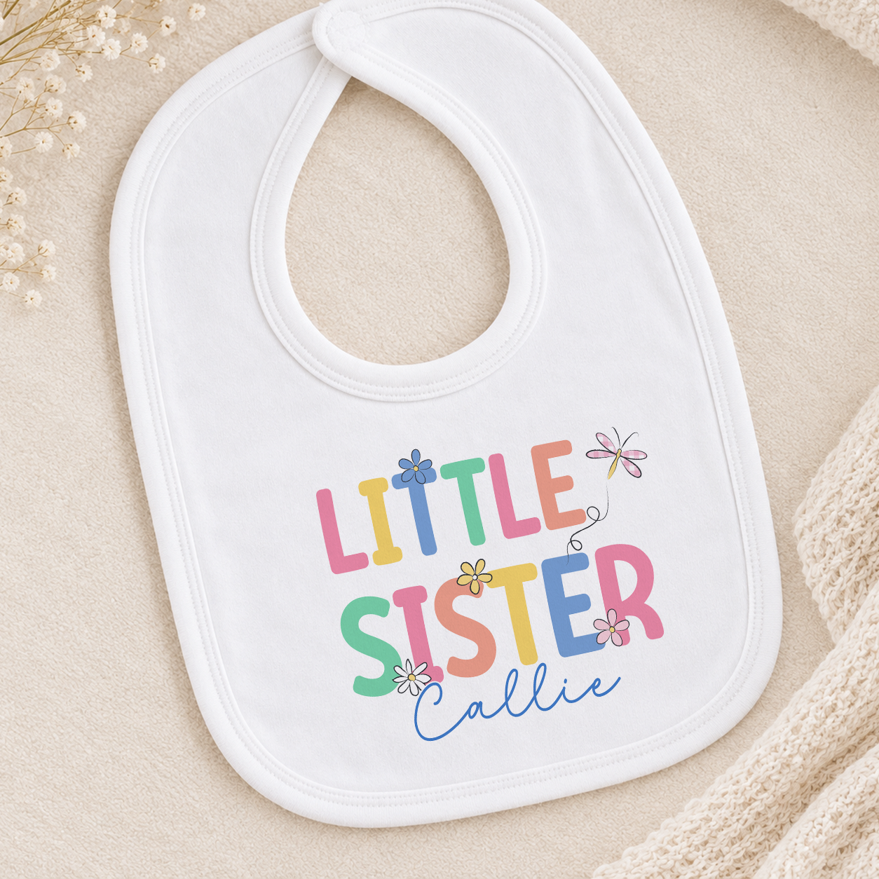 A white baby bib with the text "Little Sister" printed on it, along with colorful flower illustrations.