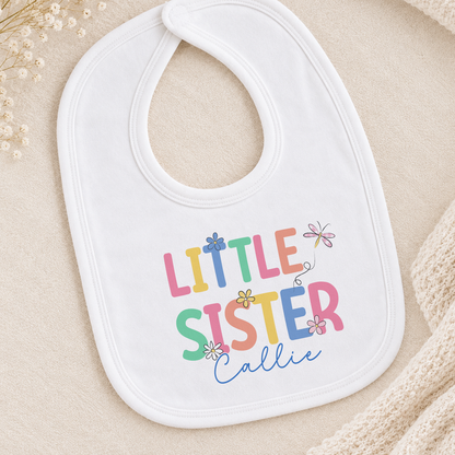 A white baby bib with the text "Little Sister" printed on it, along with colorful flower illustrations.