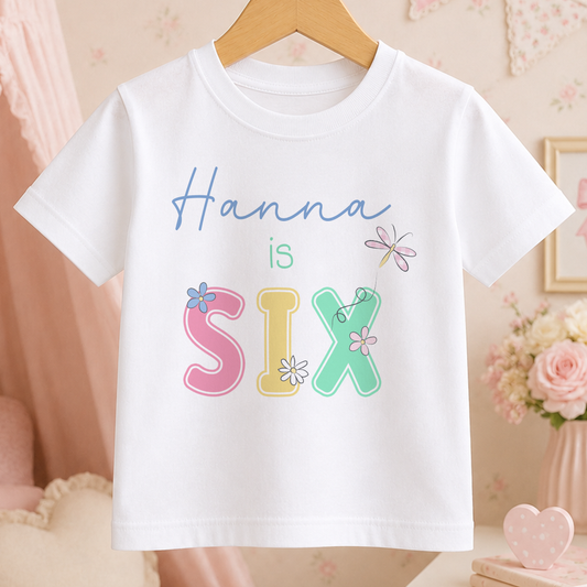 A white t-shirt with the text "Hannah is Six" printed on it, surrounded by colorful floral designs.