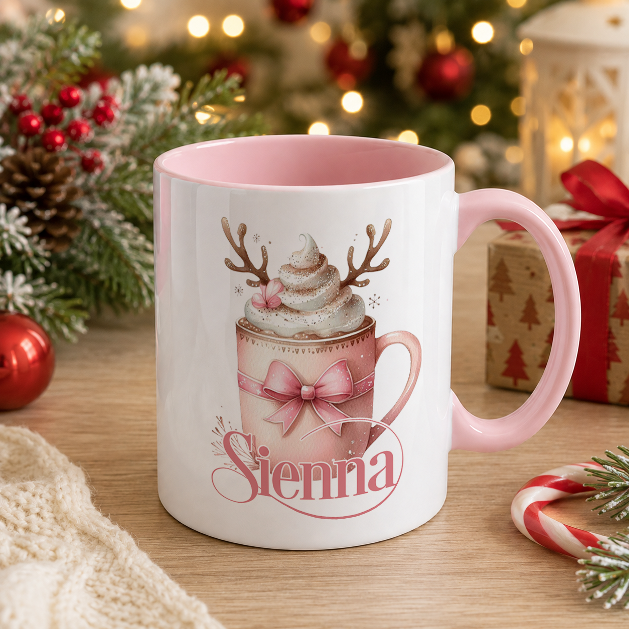 A white mug with a pink interior and a design featuring a hot chocolate drink with whipped cream and antlers, along with the name "Senna" written on it. The mug is placed on a wooden surface, surrounded by Christmas decorations such as a pine tree, ornaments, and a gift box.