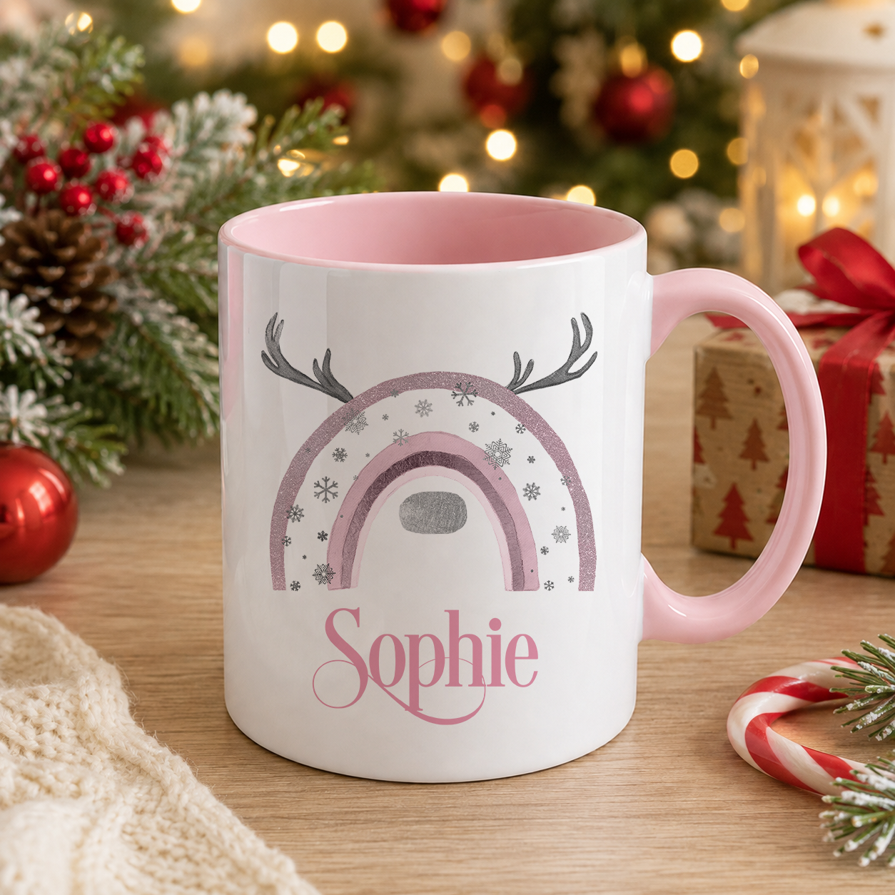 A white mug with a pink interior and a rainbow design, featuring the name "Sophie" written on it. The mug is placed on a wooden surface, surrounded by Christmas decorations such as pine cones, ornaments, and a small gift box.