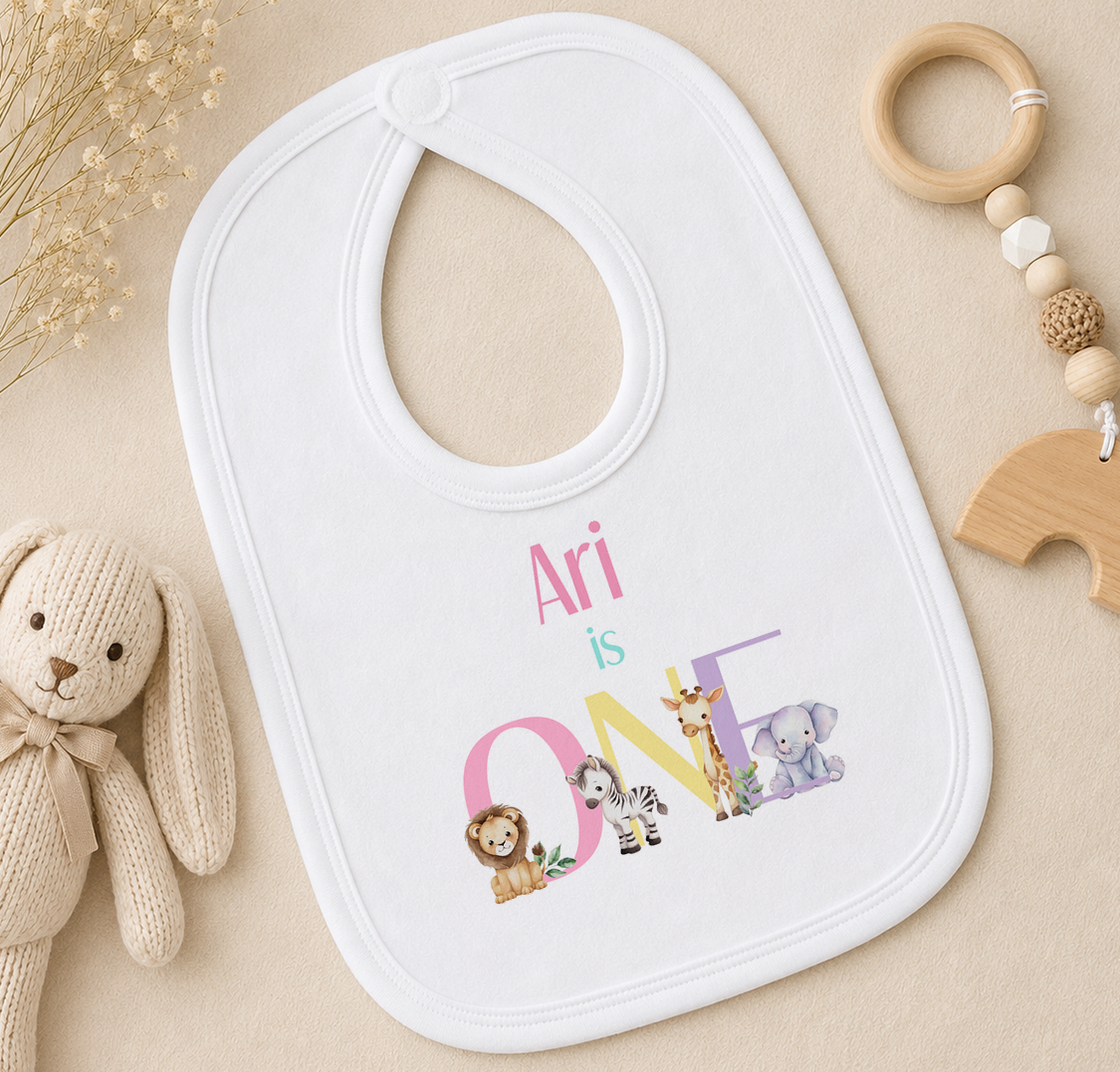 A white baby bib with the text "Ari is ONE" printed on it, surrounded by various baby items such as a wooden rattle, a stuffed animal, and a stuffed rabbit.