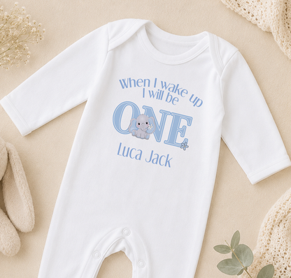 A white onesie with a blue elephant graphic and text that says "When I wake up I will be ONE Luca Jack" is displayed on a beige surface.