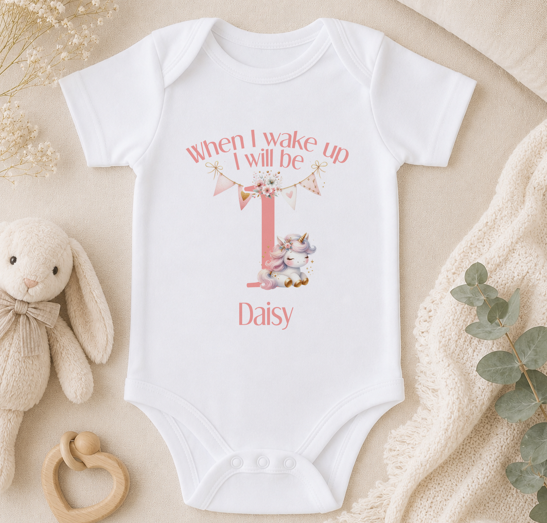 A white onesie with a personalized message and a unicorn graphic, placed on a beige surface with a stuffed animal and greenery.