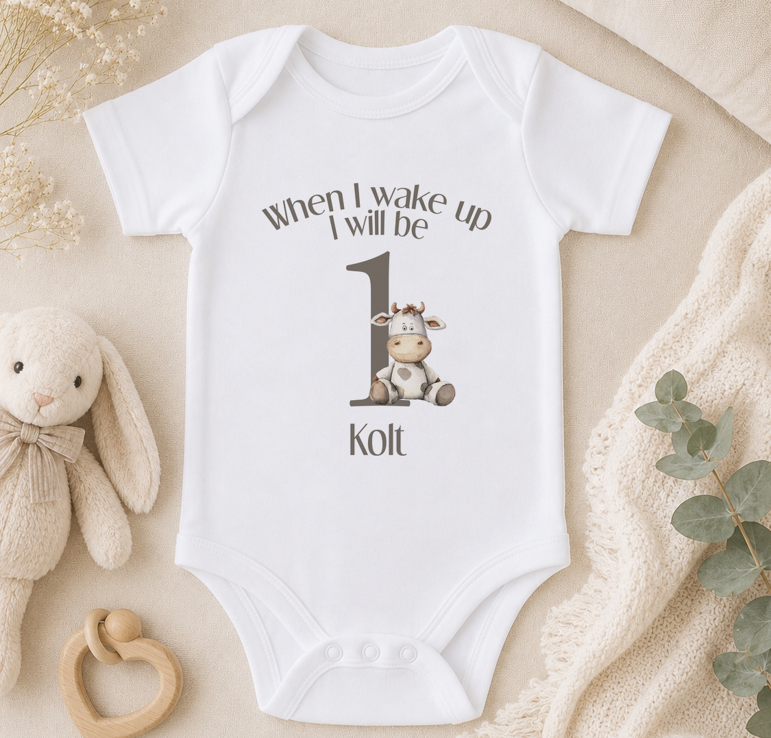 A white baby onesie with a cow graphic and the text "When I wake up I will be 1" printed on it, along with the name "Kolt". The onesie is placed on a beige blanket, and there is a stuffed animal and some greenery in the background.
