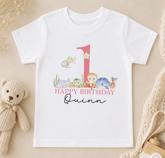 A white t-shirt with a pink number one and a "Happy Birthday Quinn" message printed on it, along with various animal illustrations.