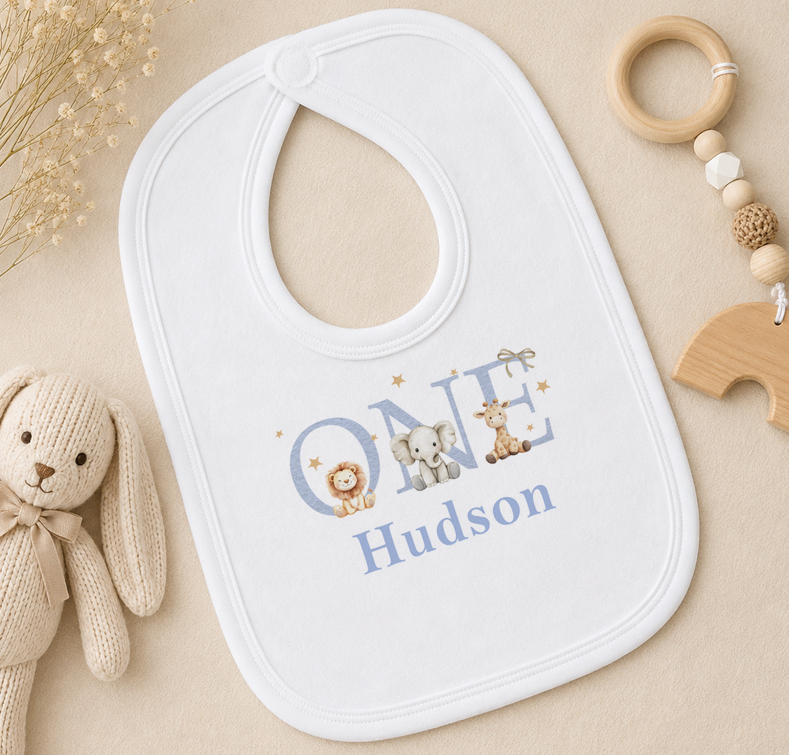 A white baby bib with the name "Hudson" printed on it, surrounded by various baby items such as a wooden rattle and a stuffed animal.
