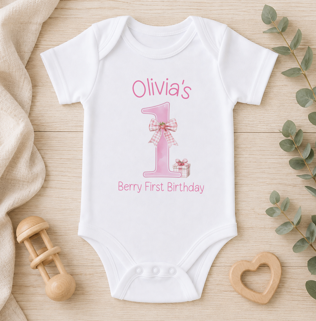 A white onesie with a pink number one and the text "Olivia's Berry First Birthday" printed on it, along with a wooden binky and a wooden heart-shaped object.