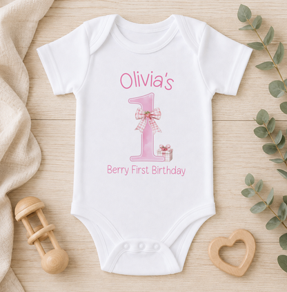 A white onesie with a pink number one and the text "Olivia's Berry First Birthday" printed on it, along with a wooden binky and a wooden heart-shaped object.