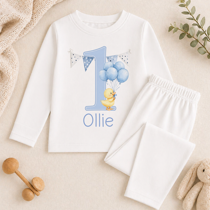A white long-sleeved shirt with a blue number one and a yellow duck, along with a pair of white pants, are displayed on a beige surface. There are also some stuffed animals and a plant in the background.
