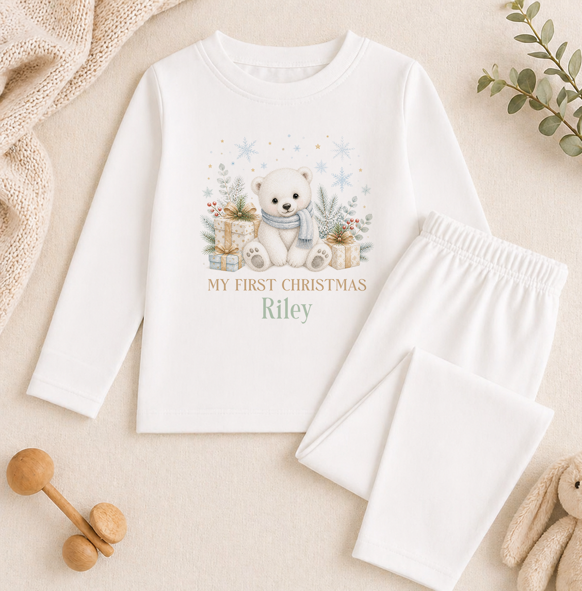 A white pajama set with a teddy bear graphic and the text "My first Christmas Riley" printed on it, along with a pair of white pants.