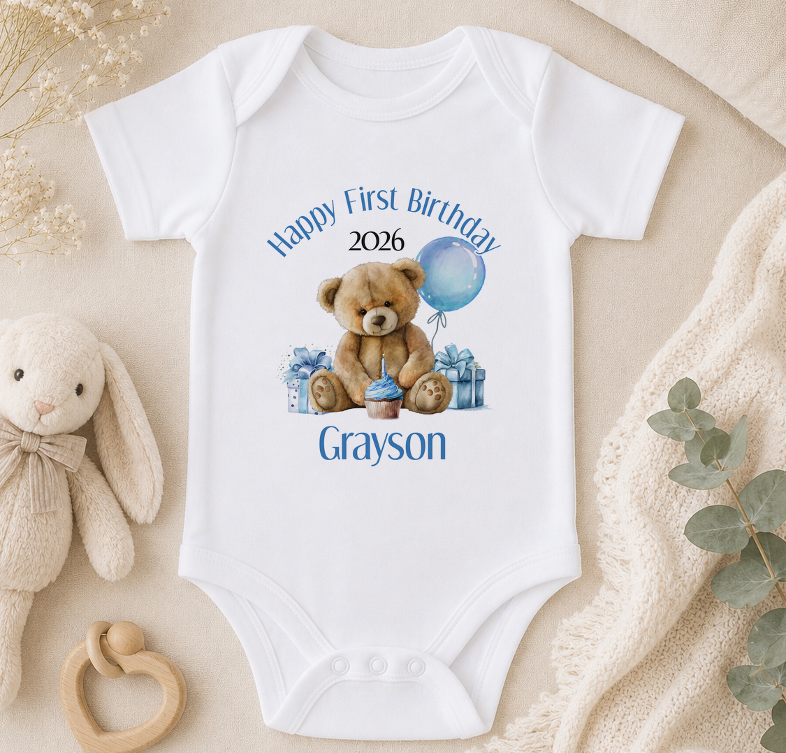A white onesie with a teddy bear and a blue balloon, along with the text "Happy First Birthday Grayson" printed on it. The onesie is placed on a beige surface, surrounded by a stuffed bunny, a wooden toy, and some greenery.