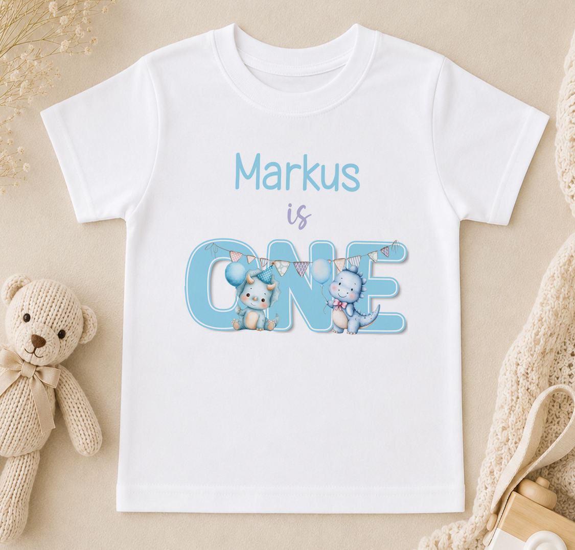A white t-shirt with a personalized text and illustrations, including a teddy bear and two cartoon animals, is displayed on a beige surface.