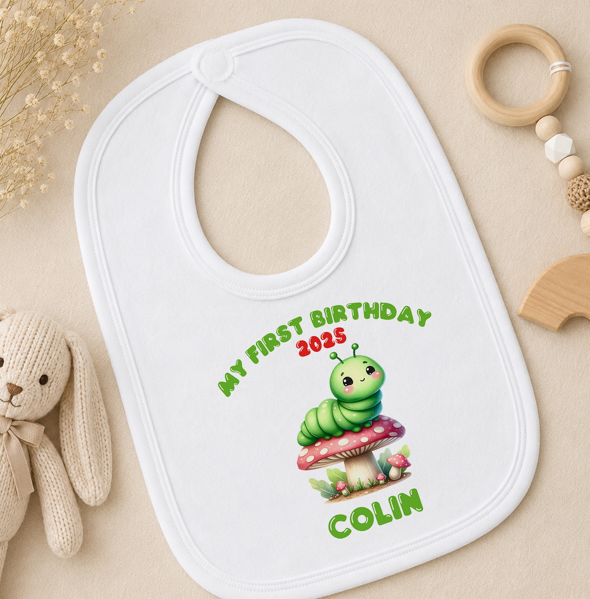 A white baby bib with a caterpillar design and the text "My first birthday 2025" printed on it, along with the name "COLIN" below the caterpillar.