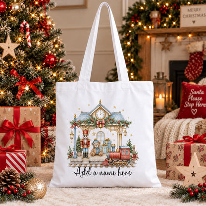 White cotton tote bag printed with a watercolour Christmas station scene with a steam train, nutcracker and teddy, above a name.