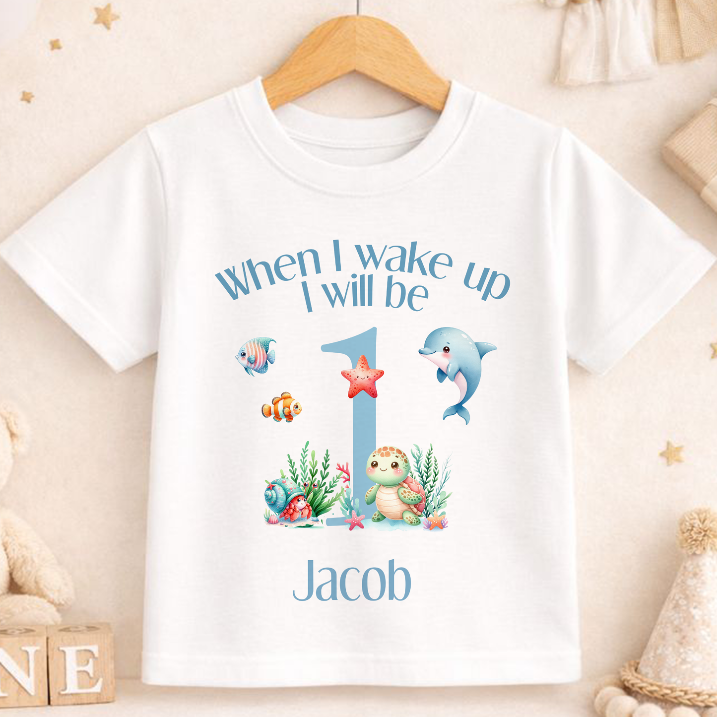 A white t-shirt with a personalized design featuring a dolphin, starfish, and other sea creatures, along with the text "When I wake up I will be Jacob" printed on it.