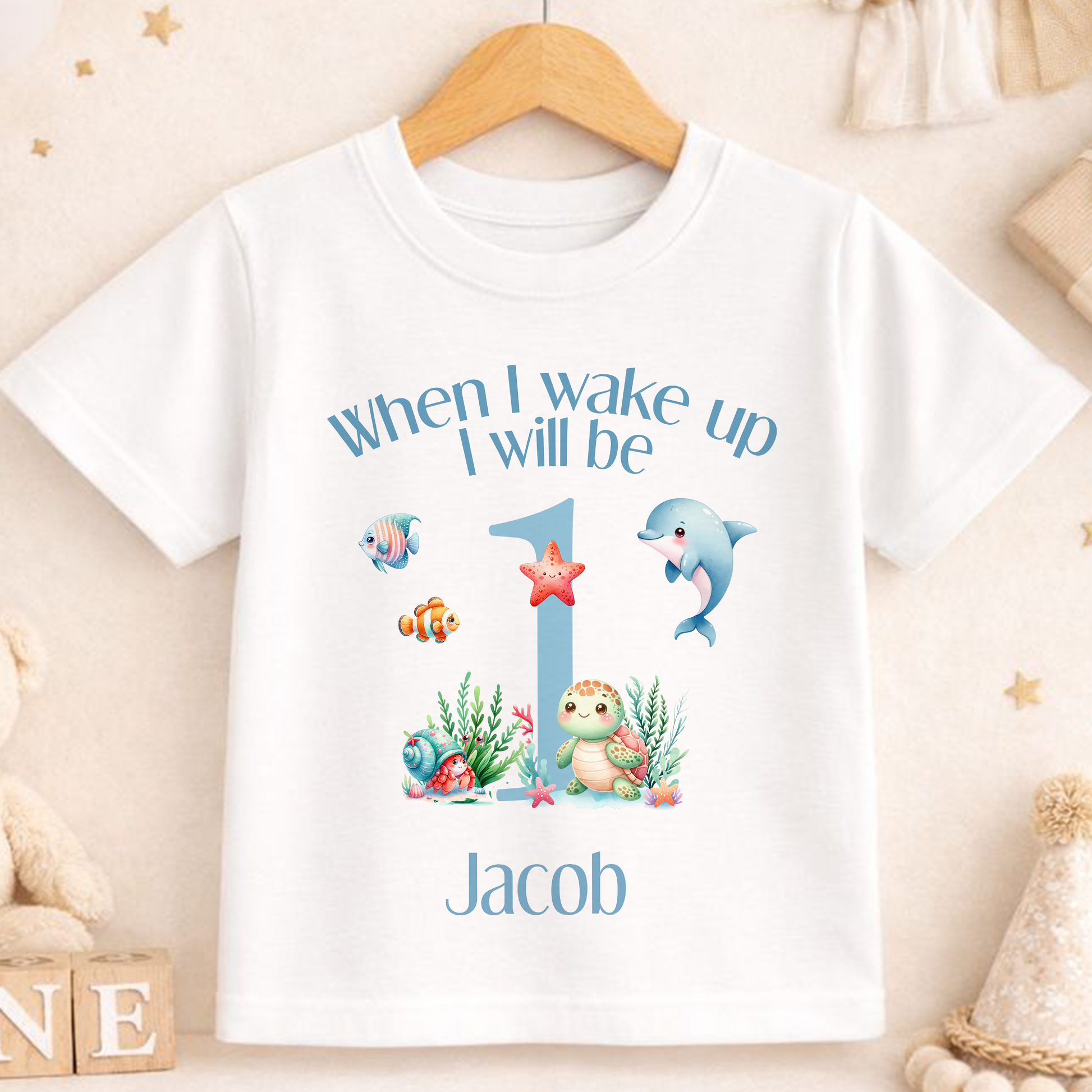 A white t-shirt with a personalized design featuring a dolphin, starfish, and other sea creatures, along with the text "When I wake up I will be Jacob" printed on it.