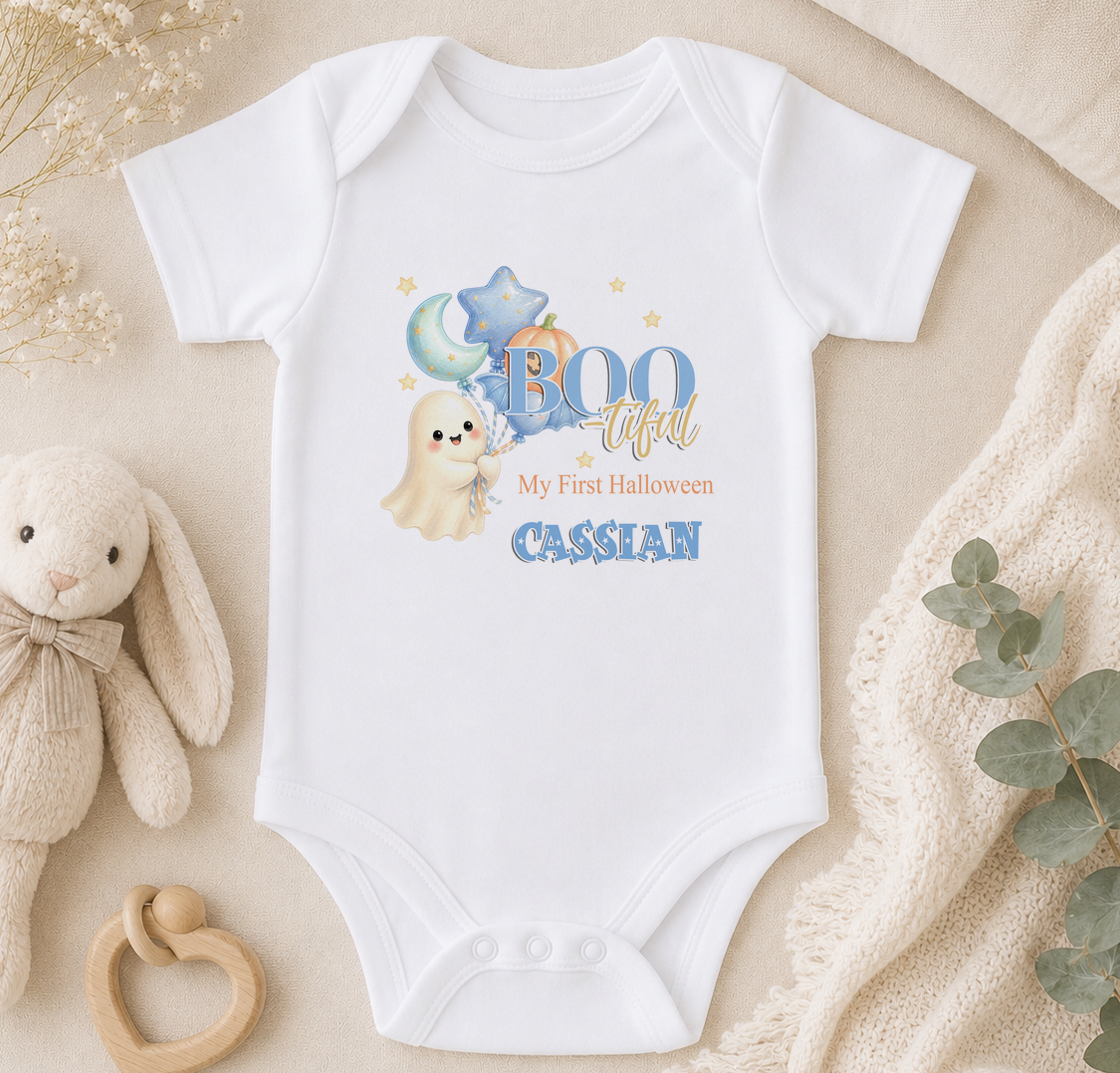 A white baby onesie with a ghost and stars design, along with the text "BOO tacular My first Halloween Cassian". The onesie is placed on a beige surface, surrounded by a stuffed bunny, a wooden bead ring, and some greenery.