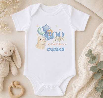 A white baby onesie with a ghost and stars design, along with the text "BOO tacular My first Halloween Cassian". The onesie is placed on a beige surface, surrounded by a stuffed bunny, a wooden bead ring, and some greenery.