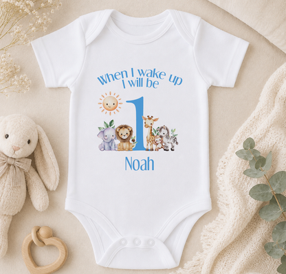 A white onesie with a cartoon animal design and the text "When I wake up I will be Noah" printed on it, along with a sun and moon graphic. The onesie is placed on a beige blanket, and there is a stuffed animal and a wooden bead ring nearby.