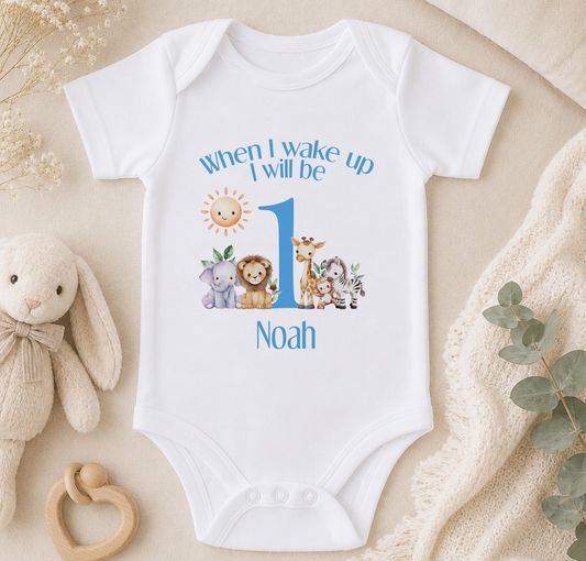 A white onesie with a cartoon animal design and the text "When I wake up I will be Noah" printed on it, along with a sun and moon graphic. The onesie is placed on a beige blanket, and there is a stuffed animal and a wooden bead ring nearby.