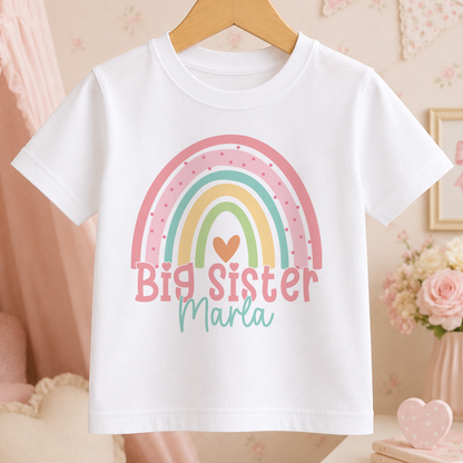 A white t-shirt with a colorful rainbow graphic and the text "Big Sister Maria" printed on it, hanging on a wooden hanger.