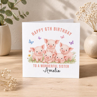 Square birthday card with five pink piglets in a flower field surrounded by butterflies, personalised Happy 6th Birthday to a Wonderful Sister Amelia, standing with its envelope.
