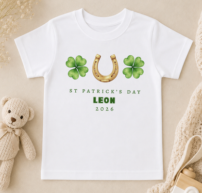 A white t-shirt with a graphic design featuring a horseshoe, four-leaf clovers, and the text "ST PATRICK'S DAY LEON 2026".
