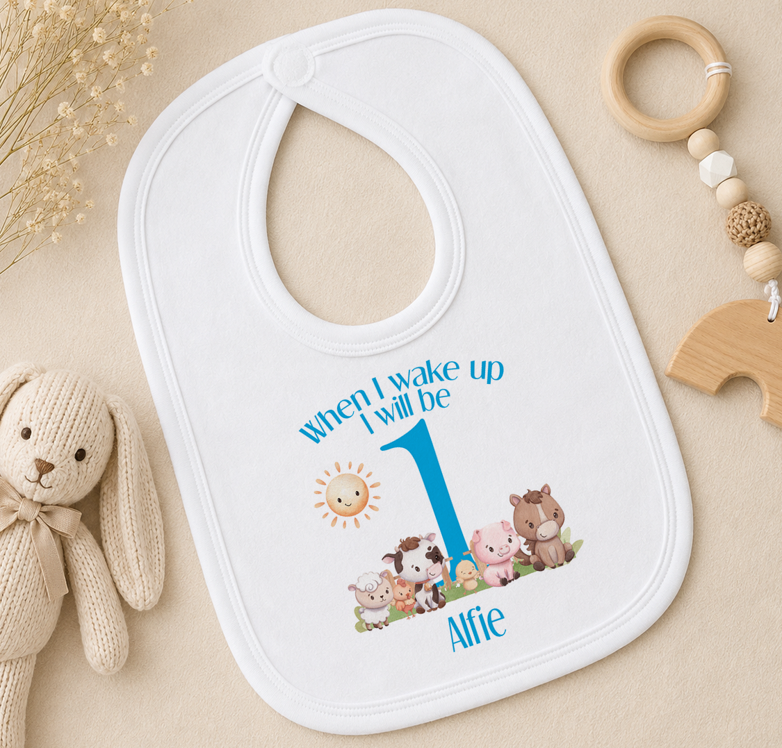 A white baby bib with a personalized message and cartoon animals printed on it, placed on a beige surface alongside a wooden toy and a wooden bead ring.