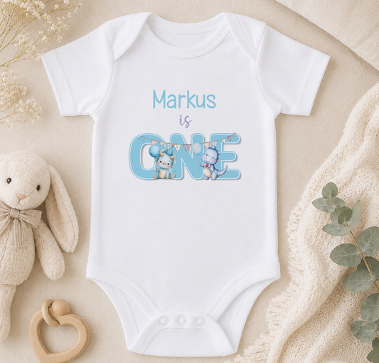 A white onesie with the text "Markus is ONE" printed on it, along with illustrations of a bunny and a cat. The onesie is placed on a beige surface, surrounded by a stuffed animal, a wooden toy, and some greenery.
