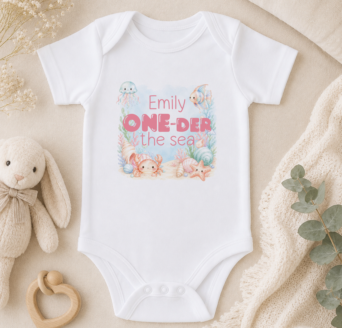 Personalised baby onesie printed with sea creatures and the phrase 'Emilly ONE-DER the sea', styled with a soft toy rabbit