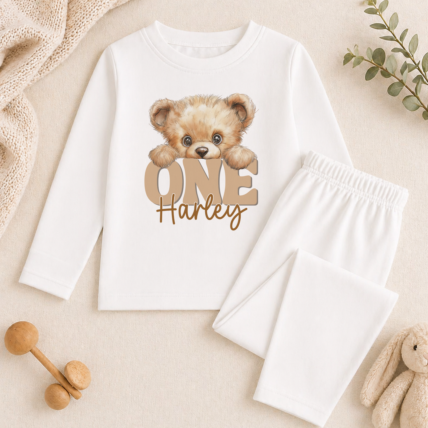 A white long-sleeved shirt with a teddy bear design and the text "One Harley" printed on it, along with a pair of white pants. There are also some wooden toys and a plant in the background.