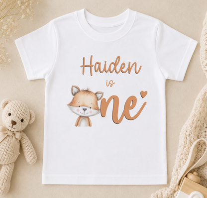 A white t-shirt with a cartoon image of a fox and the text "Haiden is one" printed on it, along with a teddy bear and other baby items.