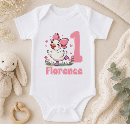 A white onesie with a cartoon duck and the name "Florence" printed on it, along with a pink bow and the number "1". The onesie is placed on a beige surface, surrounded by a stuffed animal, a wooden bead ring, and some greenery.