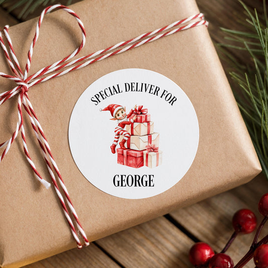 A brown gift box wrapped with red and white twine, featuring a white circular label with an illustration of an elf holding presents.