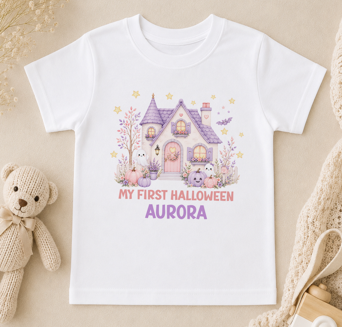 A white t-shirt with a Halloween-themed graphic design, including a house, bats, and other spooky elements, along with the text "My first Halloween Aurora".