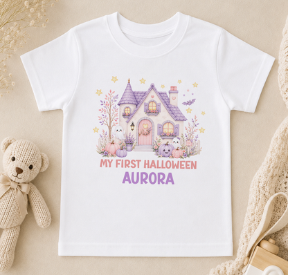 A white t-shirt with a Halloween-themed graphic design, including a house, bats, and other spooky elements, along with the text "My first Halloween Aurora".