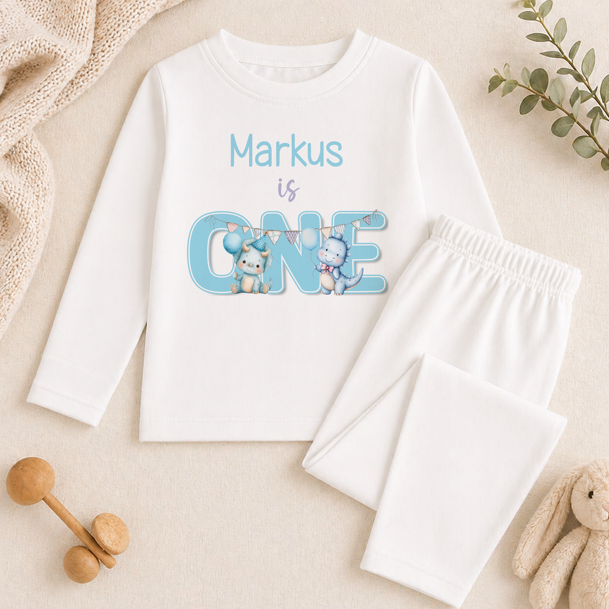 A white long-sleeved shirt with a personalized text and illustrations, along with a pair of white pants, a stuffed animal, and some decorative elements, all arranged on a beige background.