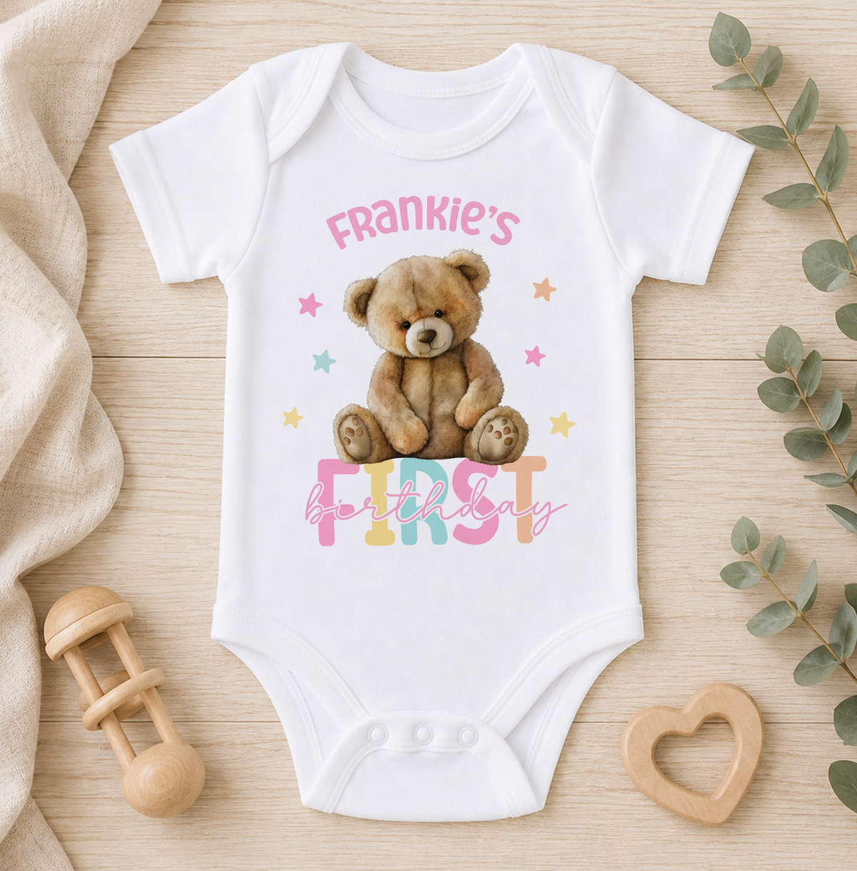 A white baby onesie with a teddy bear and the text "Frankie's first birthday" printed on it, along with a wooden baby rattle and a heart-shaped wooden toy.