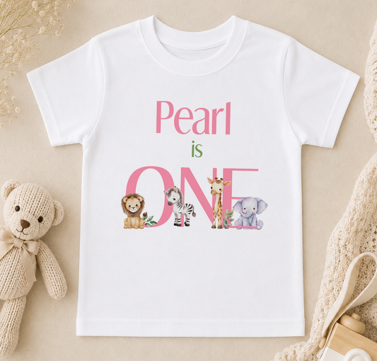 A white t-shirt with the text "Pearl is ONE" printed on it, along with illustrations of a lion, zebra, giraffe, and elephant.
