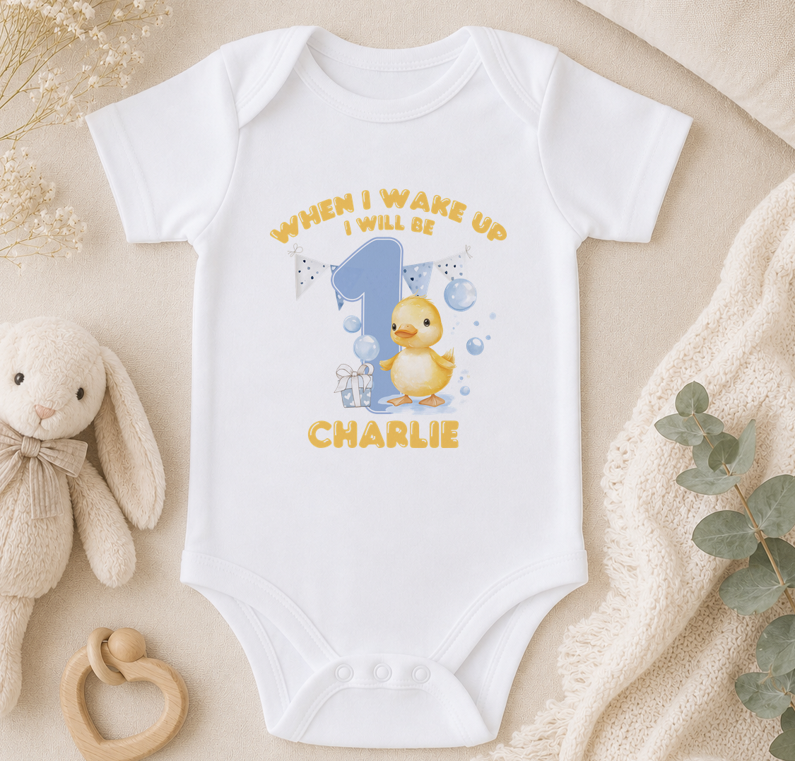 A white onesie with a yellow duck and the text "WHEN I WAKE UP I WILL BE CHARLIE" printed on it, along with a small stuffed animal and some decorative elements.