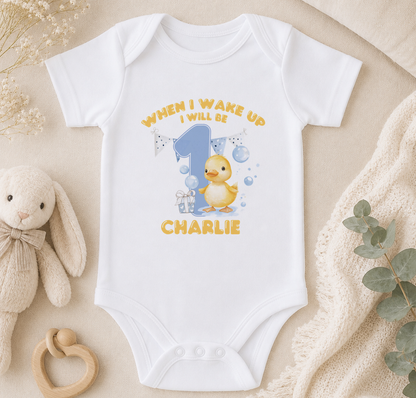 A white onesie with a yellow duck and the text "WHEN I WAKE UP I WILL BE CHARLIE" printed on it, along with a small stuffed animal and some decorative elements.