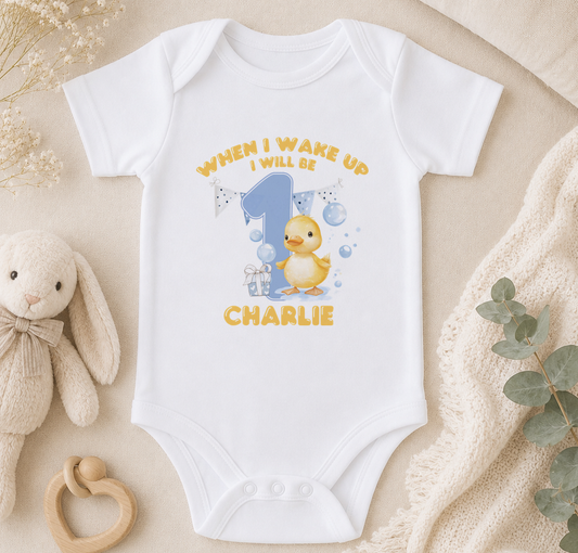 A white onesie with a yellow duck and the text "WHEN I WAKE UP I WILL BE CHARLIE" printed on it, along with a small stuffed animal and some decorative elements.