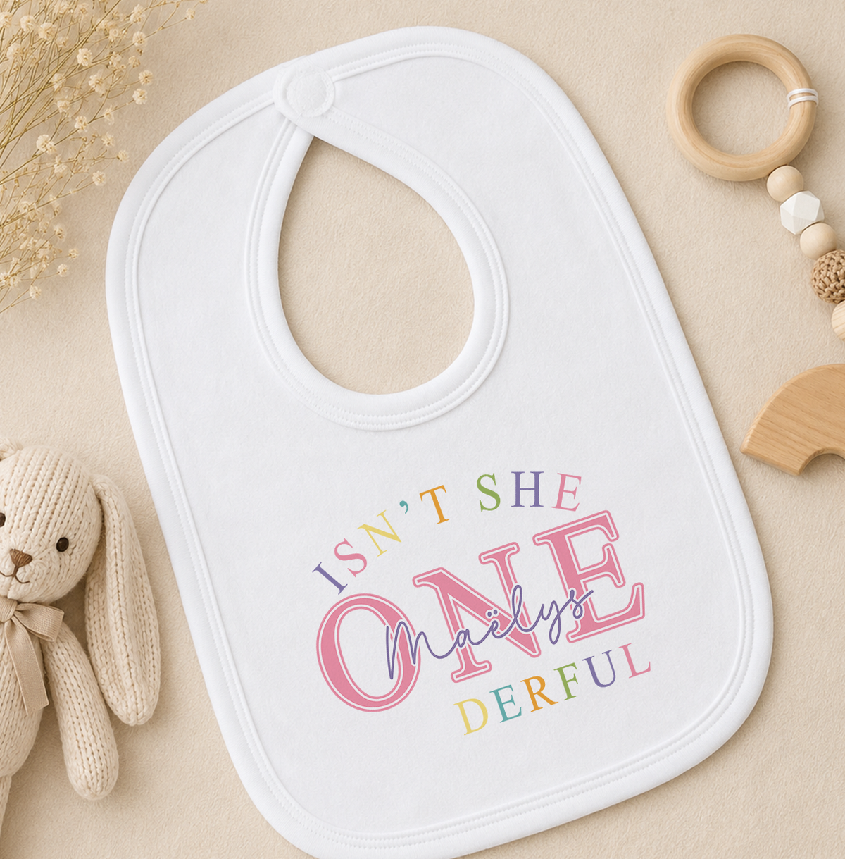 White baby bib printed with a colourful 'Isn't she ONE-derful' design, styled with a soft toy and baby toys
