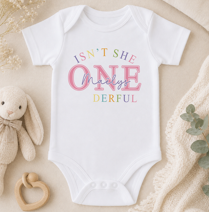 White baby onesie personalised with the phrase 'Isn't she ONE-derful' and the child's name, styled with a wooden bib and a soft toy