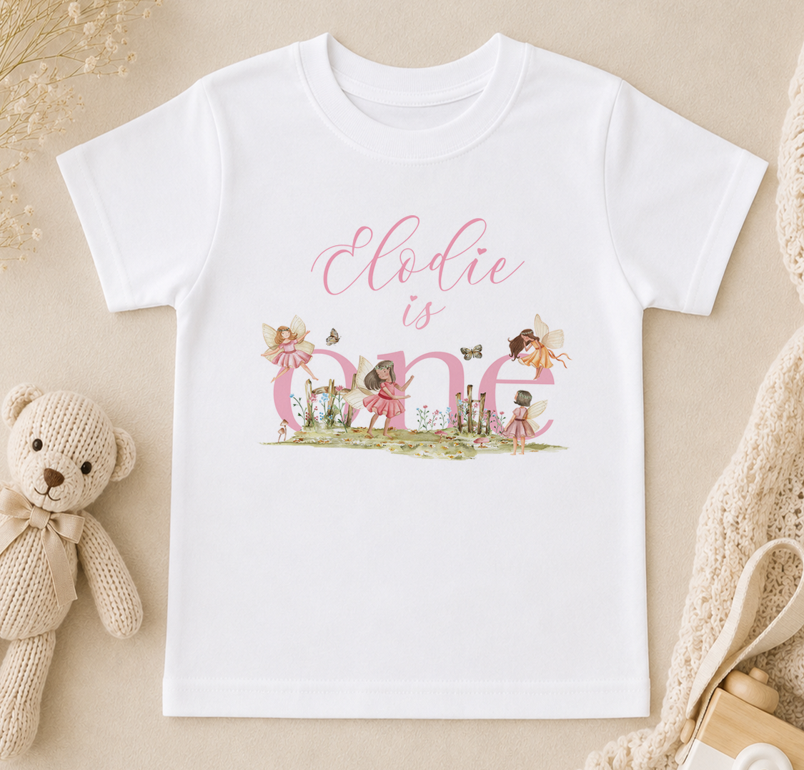 A white t-shirt with a personalized text and illustrations, including a teddy bear, fairy characters, and a garden scene, is displayed on a beige surface.