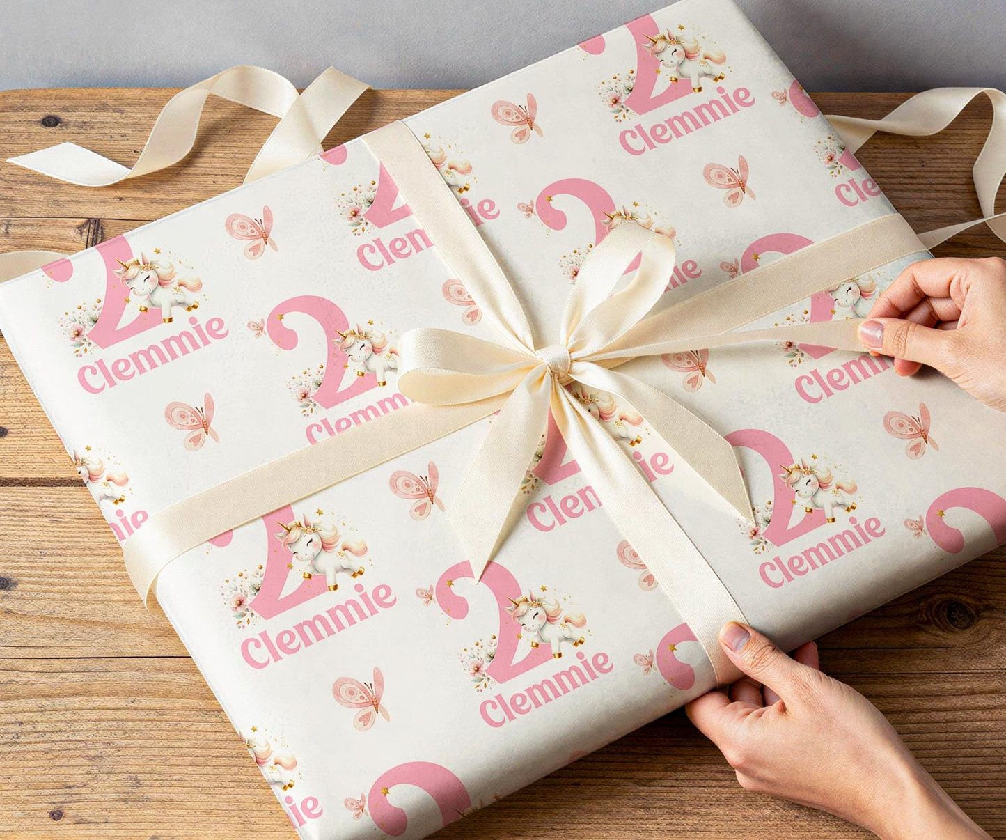 Hands wrapping a gift with white ribbon in personalised pink unicorn wrapping paper printed with a custom name