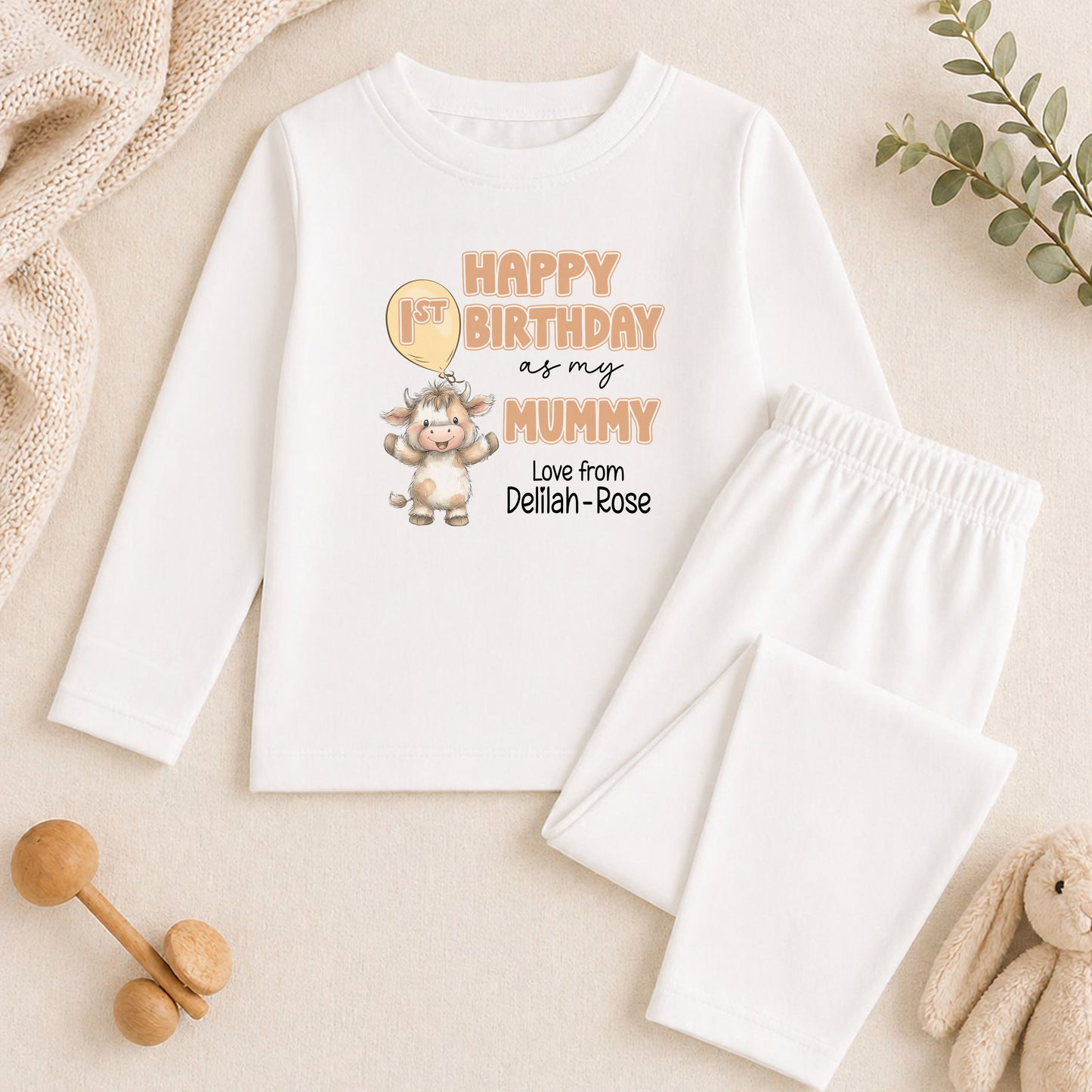 A white long-sleeved shirt with a birthday message and a cartoon image of a baby cow, along with a pair of white pants and a stuffed animal.