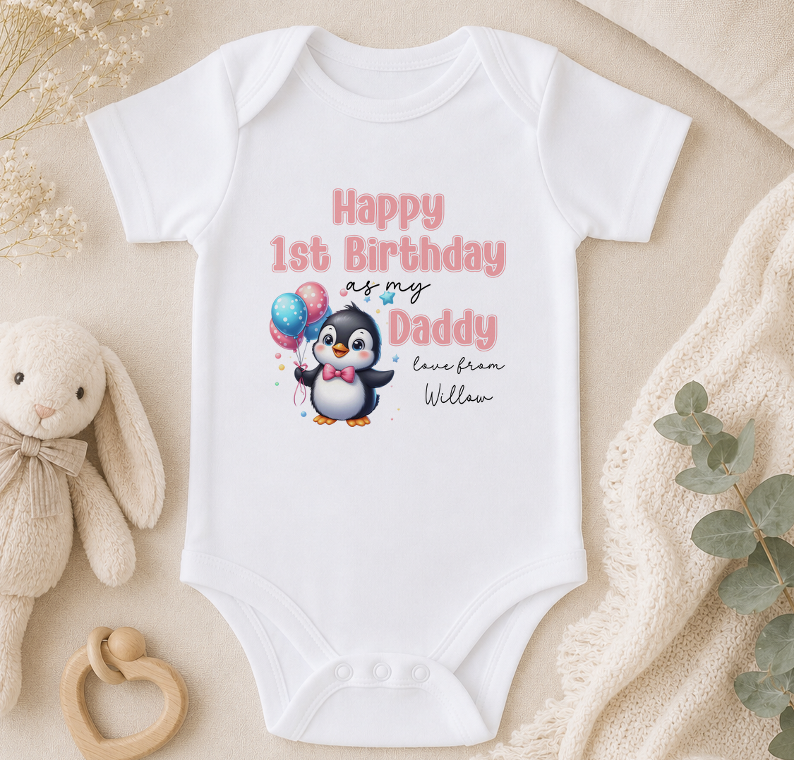 A white onesie with a penguin graphic and text that reads "Happy 1st Birthday as my Daddy love from Willow" is displayed on a beige surface, accompanied by a stuffed animal and some greenery.