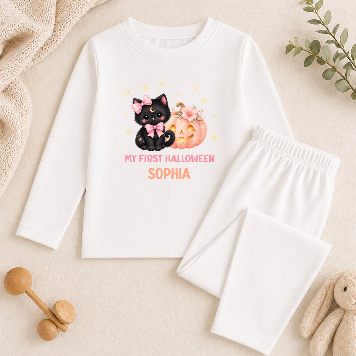 A white long-sleeved shirt with a Halloween-themed graphic of a black cat and a pumpkin, along with the text "My first Halloween Sophia" printed on it. The shirt is placed on a beige surface, and there are some toys and a plant nearby.